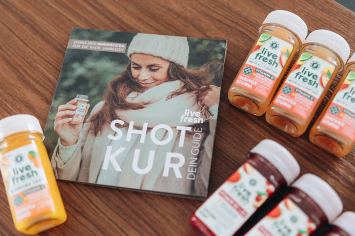 Flyer mit einer Frau und einem Shot in der Hand, sowie der weißen Aufschrift „Shot Kur – Dein Guide“ liegt auf einem Holztisch, umgeben von mehreren LiveFresh-Shots in verschiedenen Geschmacksrichtungen.