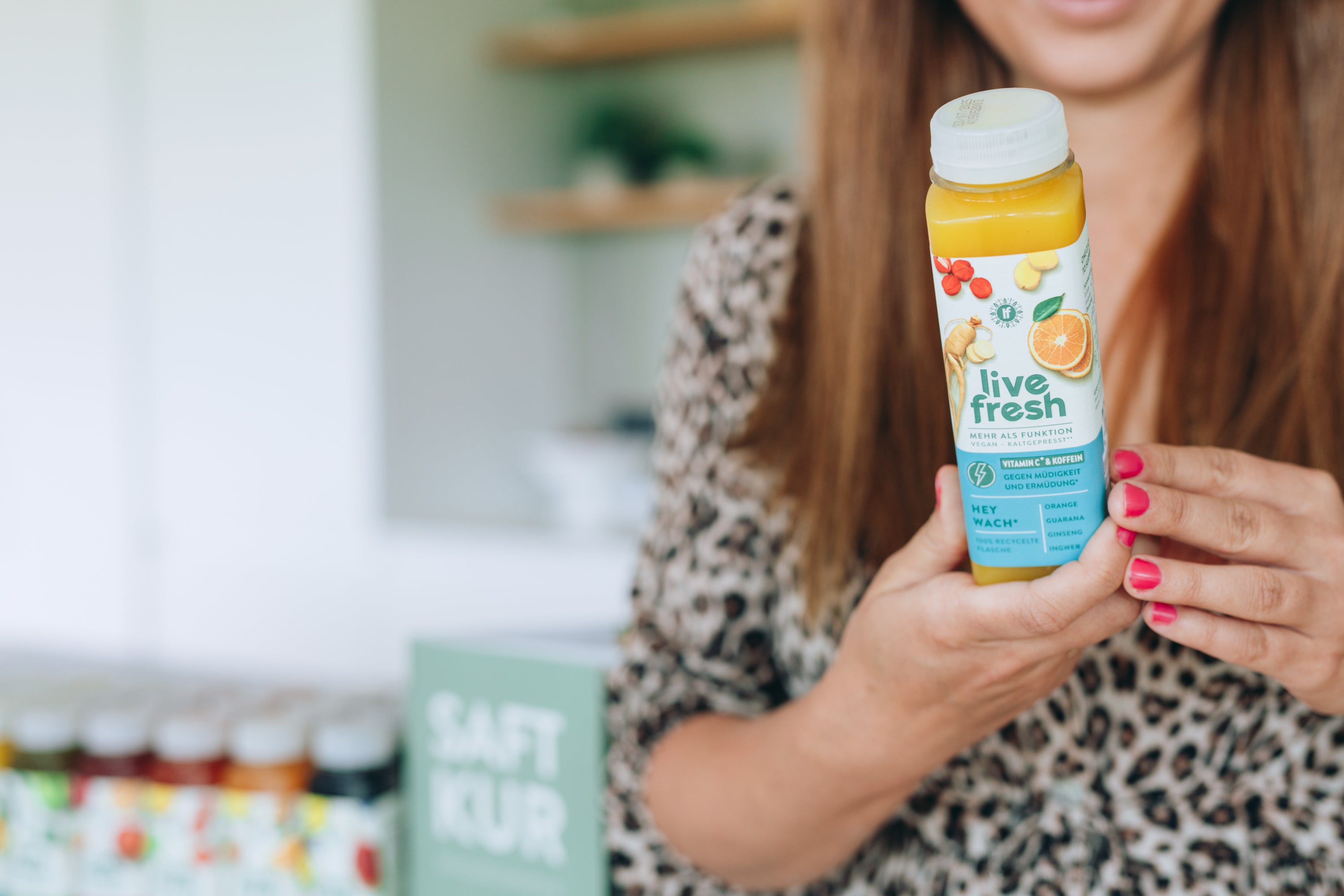 Frau mit langen Haaren und Leoparden-Top zeigt eine geschlossene Flasche „Live Fresh Hey Wach“ mit Orangen, Ingwer und Guarana.