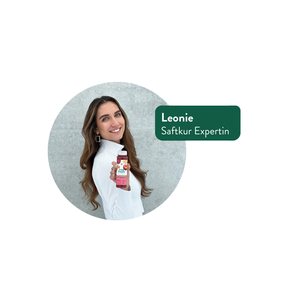 Eine lächelnde Frau mit langen braunen Haaren und weißem Oberteil hält eine Flasche LiveFresh Saft in der Hand. Neben ihr steht ein grünes Namensschild mit der Aufschrift „Leonie – Saftkur Expertin“. Der Hintergrund ist eine graue Wand.