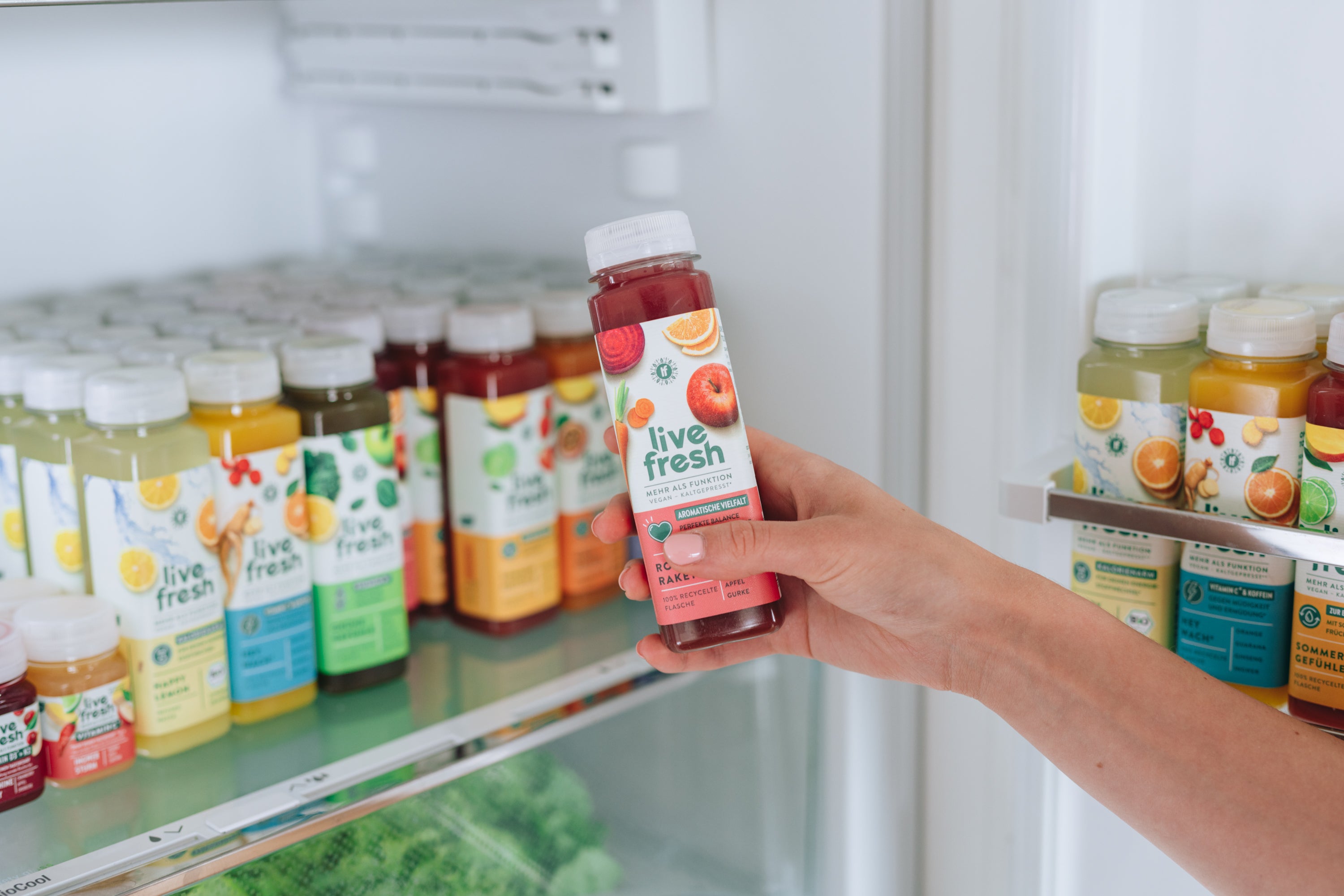 Hand greift in einen Kühlschrank und hält eine Flasche „Live Fresh“ Rote-Rakete-Saft mit Apfel und roter Bete auf dem Etikett.