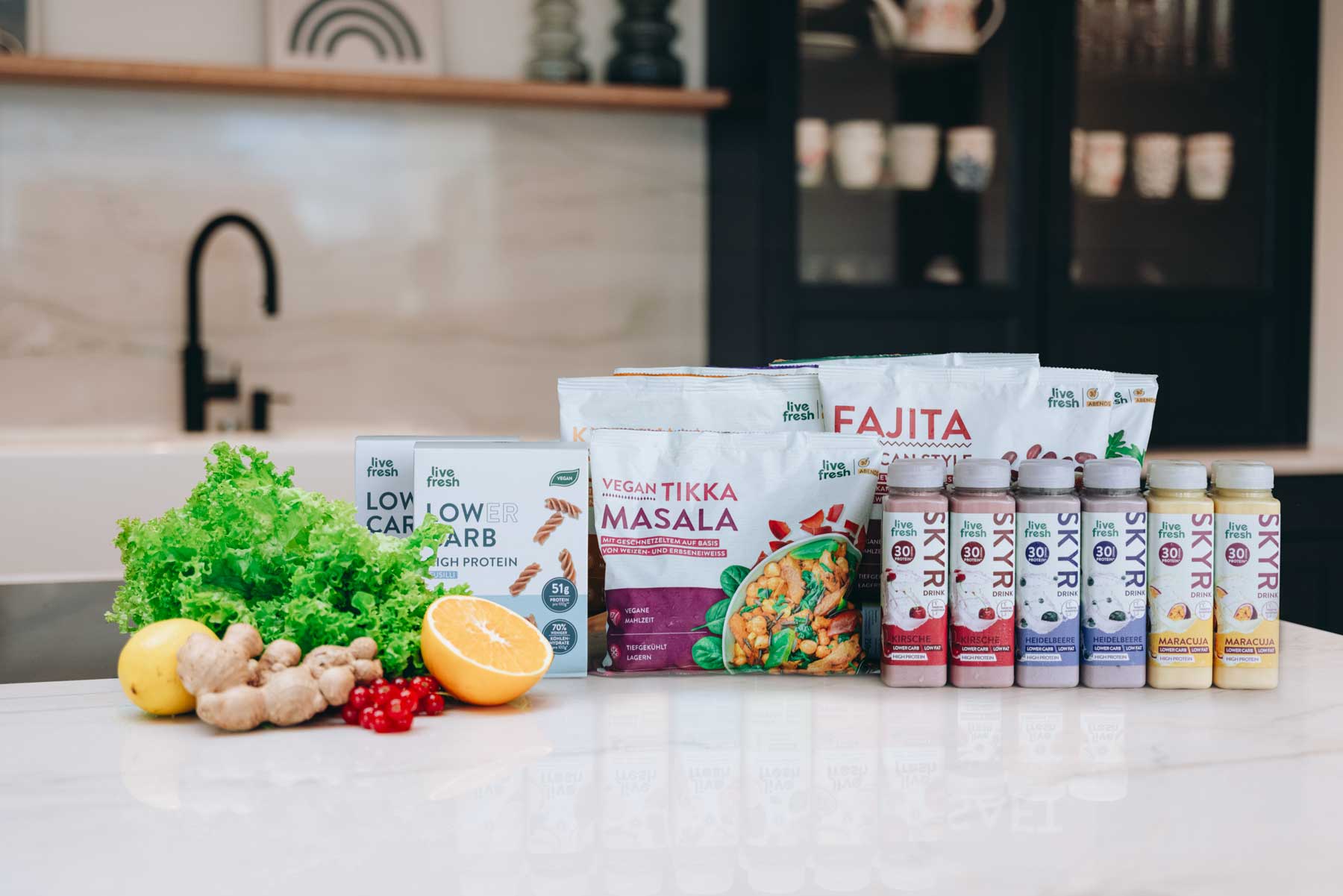 Eine Auswahl an LiveFresh Produkten auf einer Küchenarbeitsplatte, darunter Fertiggerichte wie „Vegan Tikka Masala“ und „Fajita“, Low-Carb-Nudeln sowie verschiedene Skyr-Drinks mit Kirsche, Heidelbeere und Maracuja. Salat, Ingwer, eine halbe Orange und rote Beeren liegen daneben. Im Hintergrund eine moderne Küche mit dunklen Schränken.