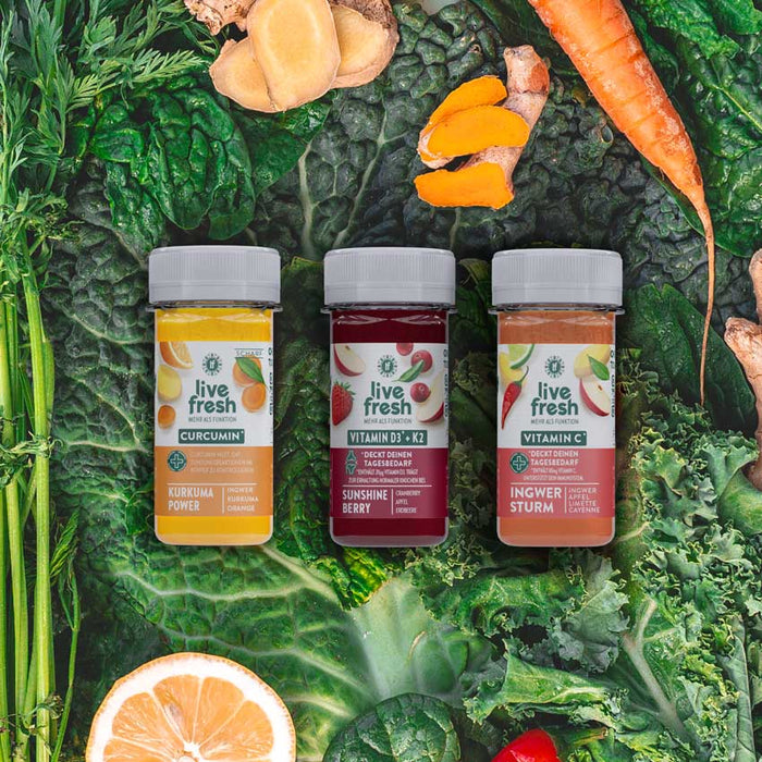 Drei „Live Fresh“-Shots in kleinen Flaschen, platziert auf einem Hintergrund aus frischem Gemüse wie Grünkohl, Karotten, Ingwer und Zitrusfrüchten. Die Sorten sind „Kurkuma Power“ (Curcumin mit Ingwer und Orange), „Sunshine Berry“ (Vitamin D3 + K2 mit Cranberry, Apfel und Acerola) und „Ingwer Sturm“ (Vitamin C mit Ingwer, Limette und Cayenne). Die natürliche, gesunde Zusammensetzung wird durch die frischen Zutaten im Hintergrund hervorgehoben.