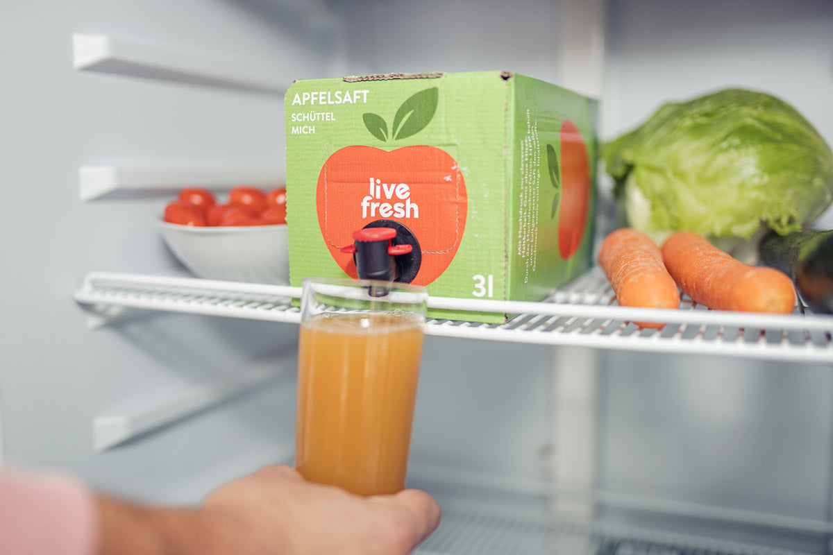 Eine grüne „LiveFresh“ Apfelsaftbox mit 3 Litern kaltgepresstem Saft im Kühlschrank. Ein volles Glas mit Apfelsaft wird von einer Hand unter dem Zapfhahn gehalten. Neben der Saftbox befinden sich Lebensmittel wie Karotten, Salat und eine Schale mit Tomaten. Auf der Box steht „Apfelsaft – Schüttel mich“ und es ist ein großer roter Apfel abgebildet.