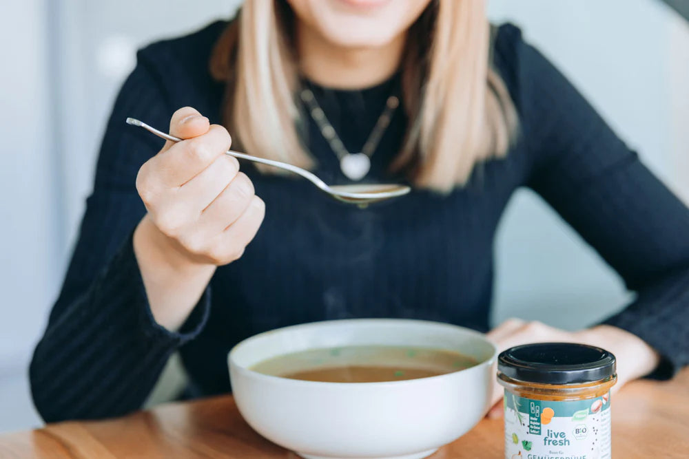 Blonde Frau in schwarzem Oberteil, deren Gesicht abgeschnitten ist, hält einen Löffel mit Gemüsebrühe über einer weißen Schale, daneben ein Glas Live Fresh Bio-Gemüsebrühe auf einem Holztisch.