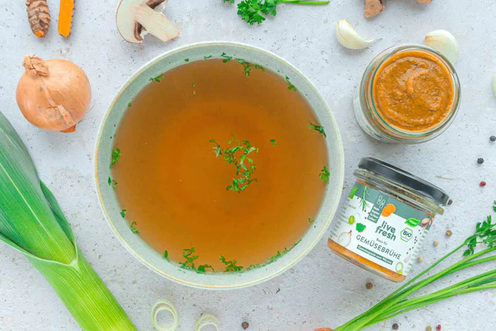 Schale mit klarer Gemüsebrühe, garniert mit Kräutern, daneben ein Glas LiveFresh Bio-Gemüsebrühe, umgeben von frischem Gemüse wie Lauch, Zwiebel und Knoblauch auf hellem Untergrund.