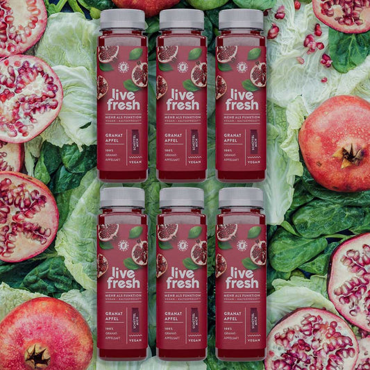Sechs „Live Fresh“ Granatapfel Säfte, arrangiert auf Blattgemüse mit aufgeschnittenen Granatäpfeln im Hintergrund.
