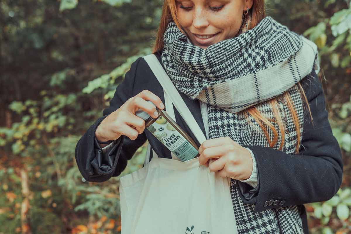Frau mit kariertem Schal und dunklem Mantel steckt eine LiveFresh-Saftflasche namens Grüne Gefühle in eine Stofftasche, umgeben von herbstlicher Waldkulisse.