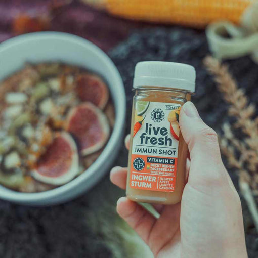 Hand hält einen Immun Shot namens Ingwer Sturm von LiveFresh mit Ingwer, Apfel, Limette und Cayenne. Im Hintergrund eine Schüssel mit Porridge, getoppt mit frischen Feigen.