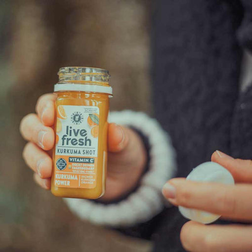 Fokus auf einen offenen Kurkuma Shot von LiveFresh, der von einer Person in der einen Hand gehalten wird, in der anderen hält sie den weißen Deckel. Die dunkle Kleidung und der Hintergrund sind verschwommen.