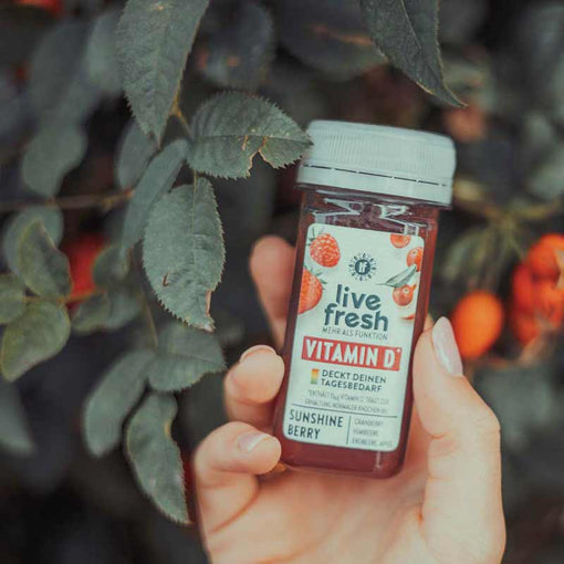 Eine Hand hält einen LiveFresh Sunshine Berry Shot mit Vitamin D vor einem Hintergrund aus grünen Blättern und roten Beeren.