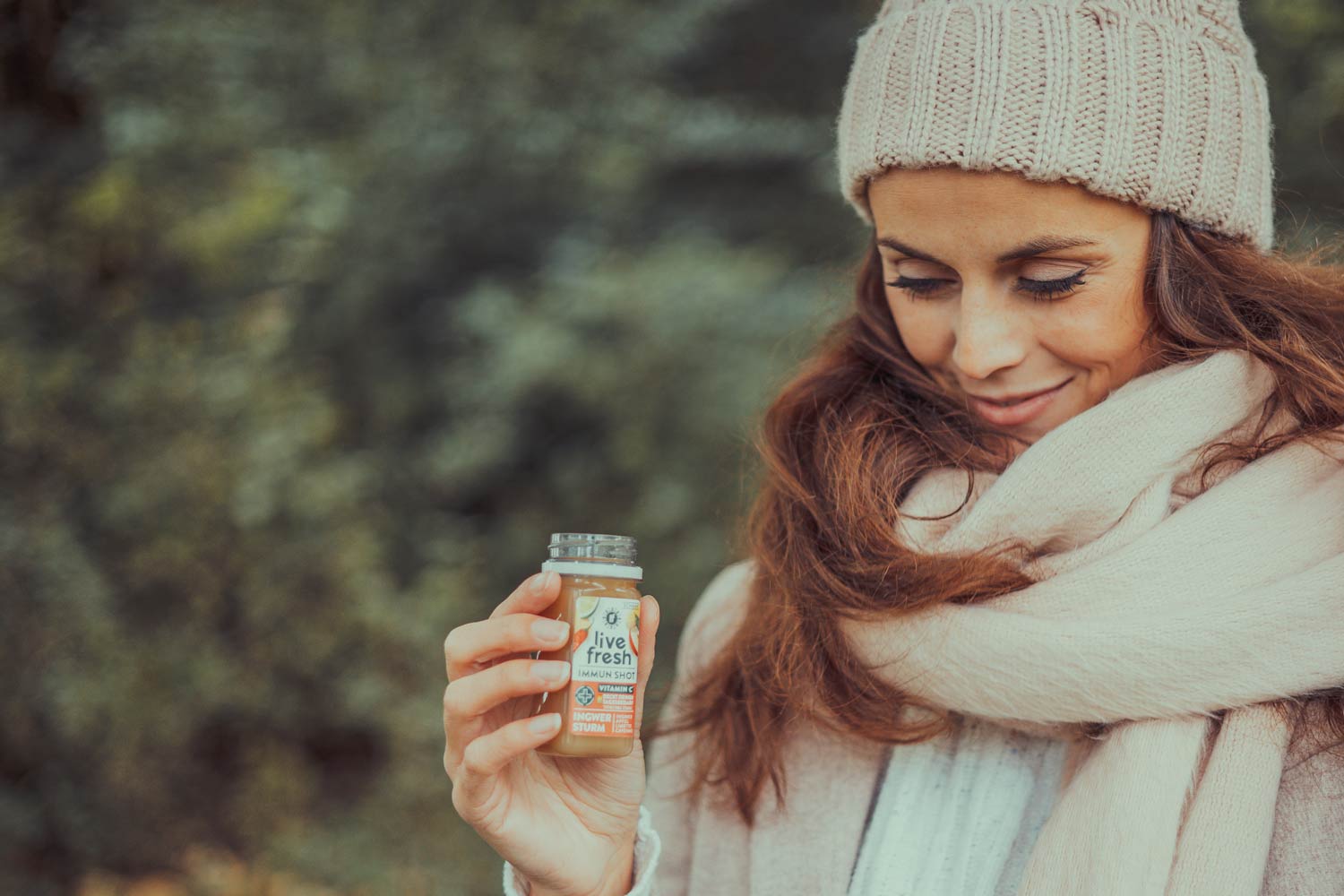 Frau mit langen, braunen Haaren, sowie beiger Strickmütze und Schal, hält einen LiveFresh Immun-Shot in der Hand und lächelt. Herbstliche Atmosphäre mit unscharfem Naturhintergrund.