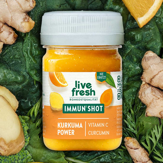 Leuchtend gelber LiveFresh Immun Shot zwischen Blattgemüse Ingwer und Orange. Auf dem Etikett steht "live fresh Rohkostqualität Immun Shot Kurkuma Power Vitamin C Curcumin".