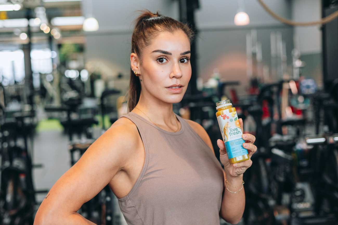 Eine sportliche Frau mit zusammengebundenem Haar steht selbstbewusst in einem Fitnessstudio und hält eine offene Flasche „Live Fresh Hey Wach“ Saft in der Hand. Sie trägt ein ärmelloses beiges Sporttop und blickt direkt in die Kamera. Im Hintergrund sind zahlreiche Fitnessgeräte unscharf zu erkennen, was die aktive, gesundheitsbewusste Atmosphäre unterstreicht.