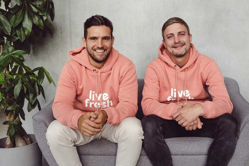 Das Bild zeigt zwei Männer in korallenfarbenen Hoodies mit dem Logo "Live Fresh", die entspannt auf einem grauen Sofa sitzen. Sie lächeln freundlich in die Kamera, was eine sympathische und zugängliche Atmosphäre vermittelt. Die große Pflanze im Hintergrund verstärkt den natürlichen, frischen Eindruck, passend zur Marke "Live Fresh".