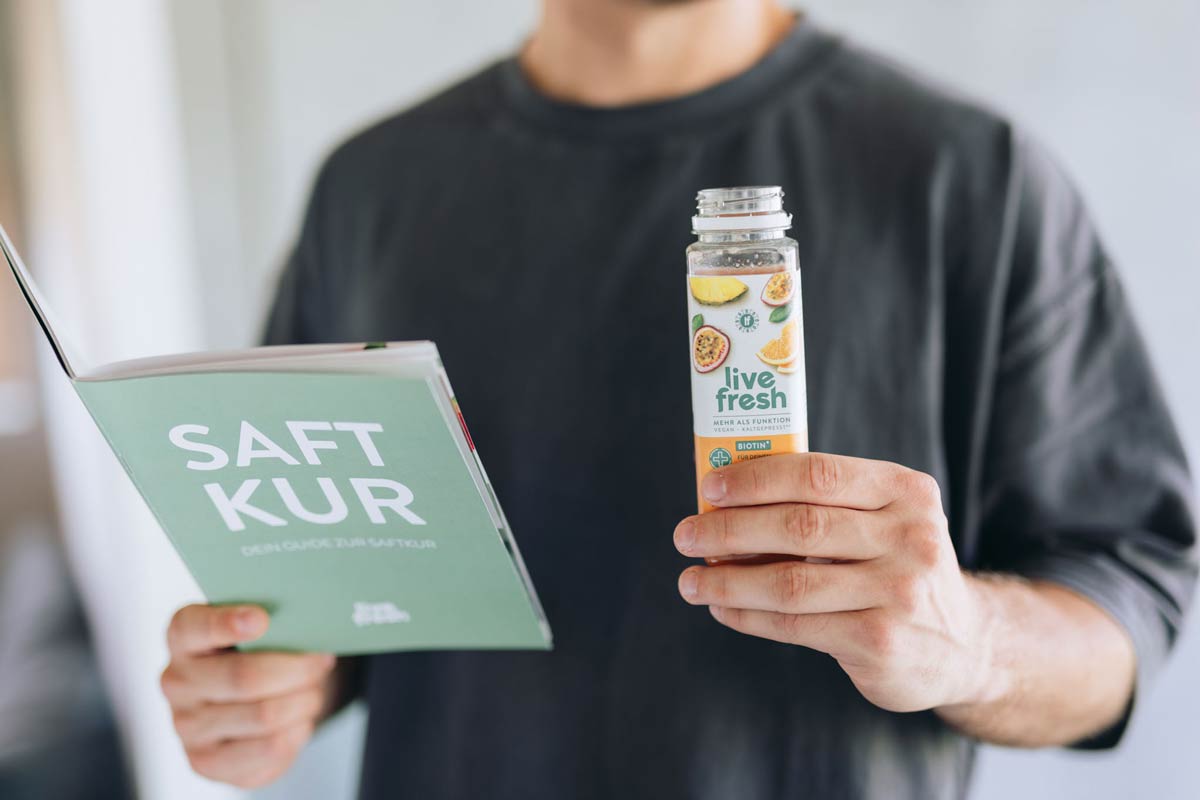 Person im grauen Shirt hält eine geöffnete Flasche LiveFresh-Saft mit Passionsfrucht, Ananas und Orange in der rechten Hand und ein grünes Booklet mit dem Titel „SAFTKUR“ in der linken Hand.