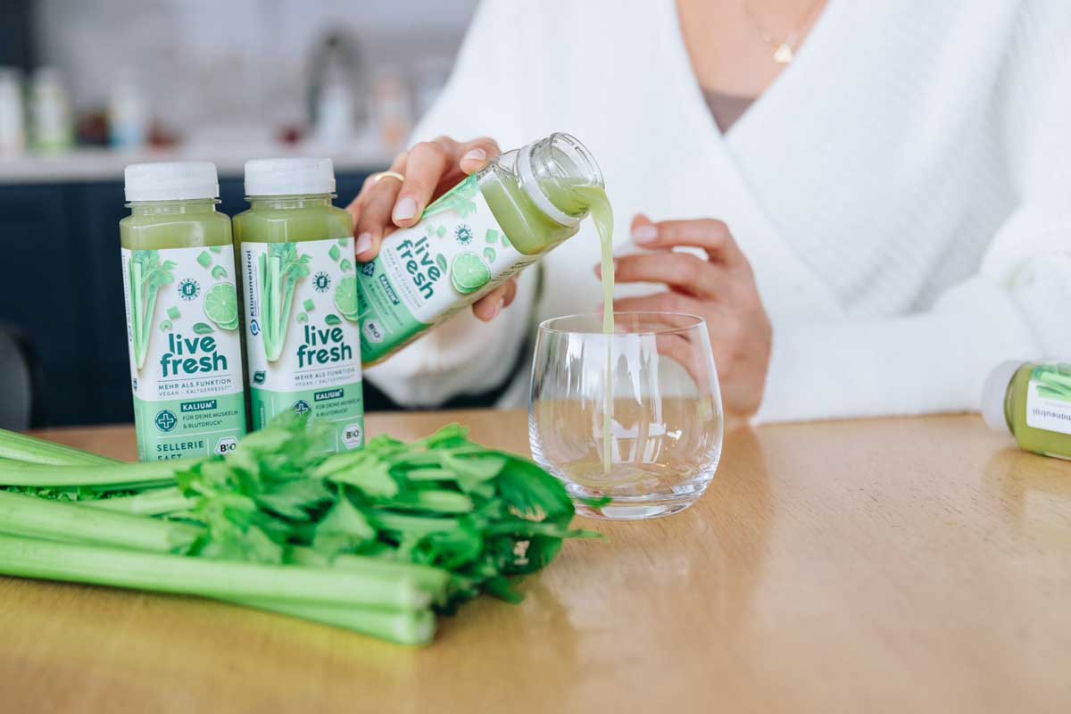 Eine Frau gießt LiveFresh Selleriesaft in ein Glas, während zwei weitere Flaschen und frische Selleriestangen auf dem Tisch liegen. Die Szene wirkt frisch und gesund, mit einem Fokus auf natürlichen, grünen Zutaten. Der helle Raum und die klare Präsentation vermitteln einen gesunden und bewussten Lebensstil.