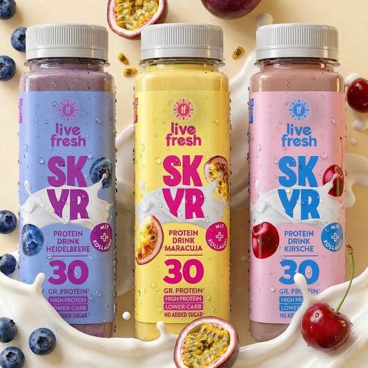 Three bottles of Skyr Protein Drink with blueberry, passion fruit and cherry stand in front of a creamy milk splash, surrounded by matching fruit. The labels say "live fresh", "SKYR", "PROTEIN DRINK BLUEBERRY", "PROTEIN DRINK MARACUJA", "PROTEIN DRINK CHERRY", "WITH COLLAGEN", "30 GR. PROTEIN", "HIGH PROTEIN", "LOWER CARB" and "NO ADDED SUGAR".