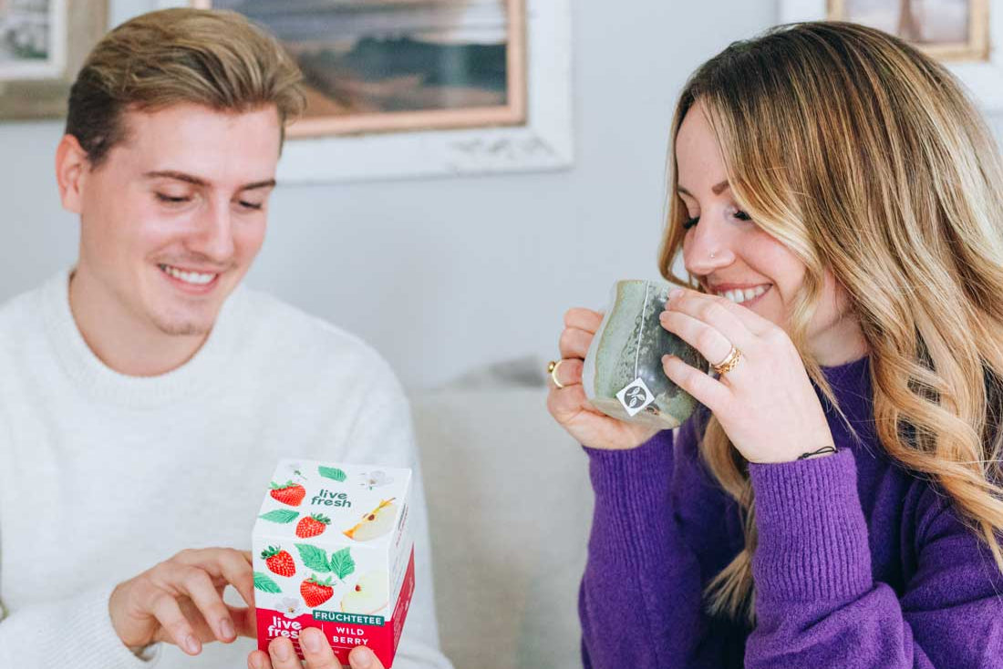 Zwei Personen auf einem Sofa – der Mann zeigt auf die Packung LiveFresh Wild Berry Tee, während die Frau lächelnd eine Teetasse in den Händen hält.