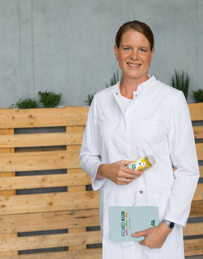 Eine lächelnde Frau mit braunen, zusammengebundenen Haaren in einem weißen Kittel hält eine Flasche Saft von LiveFresh in der einen Hand und ein Buch mit der Aufschrift