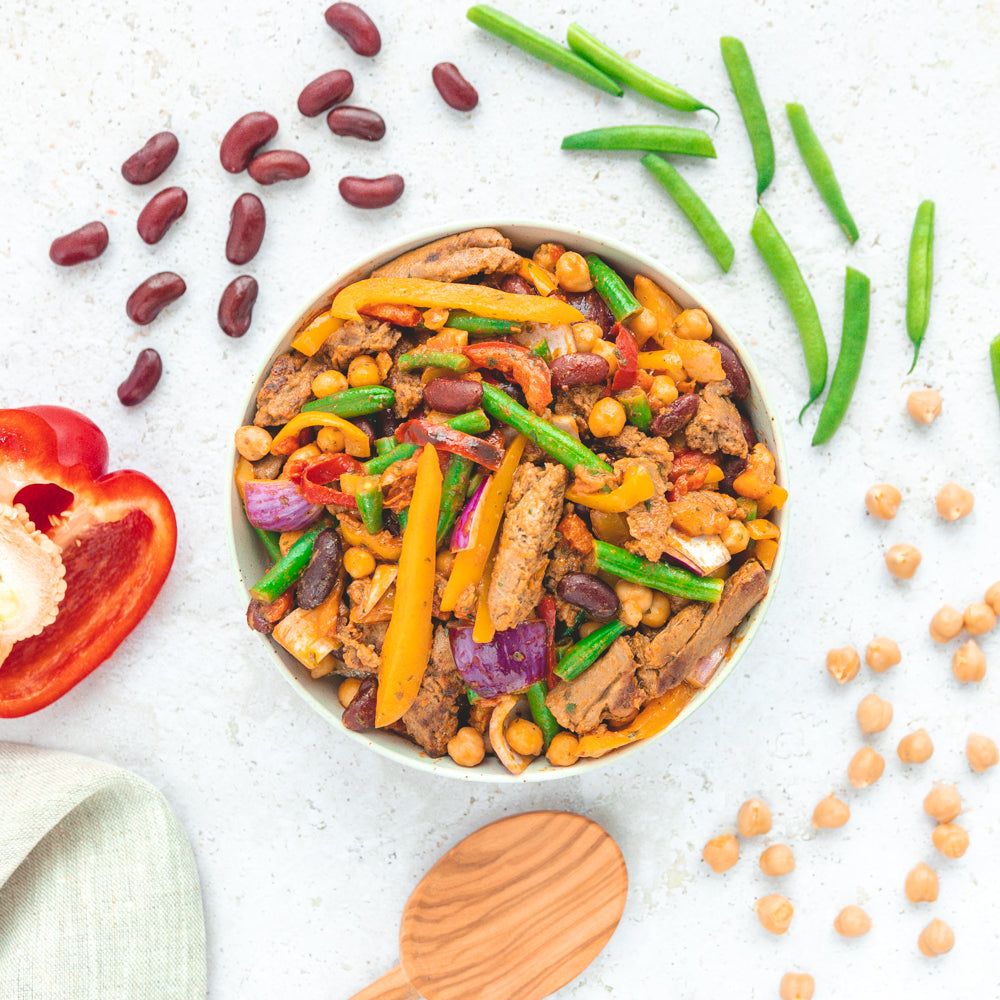 Tiefkühlgericht "Fajita Mexican Style" von LiveFresh in der Verpackung, umgeben von frischen Zutaten wie Paprika, Kidneybohnen, grünen Bohnen und Kichererbsen auf einer hellen Oberfläche. Ein Holzlöffel und ein Küchentuch befinden sich links unten.