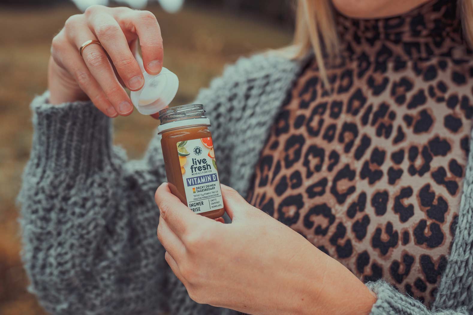 Eine Frau mit einem Leopardenmuster-Oberteil und einer grauen Strickjacke öffnet einen Shot namens Ingwer Prise von LiveFresh. Das Etikett zeigt die Zutaten wie Ingwer und Zitrusfrüchte.