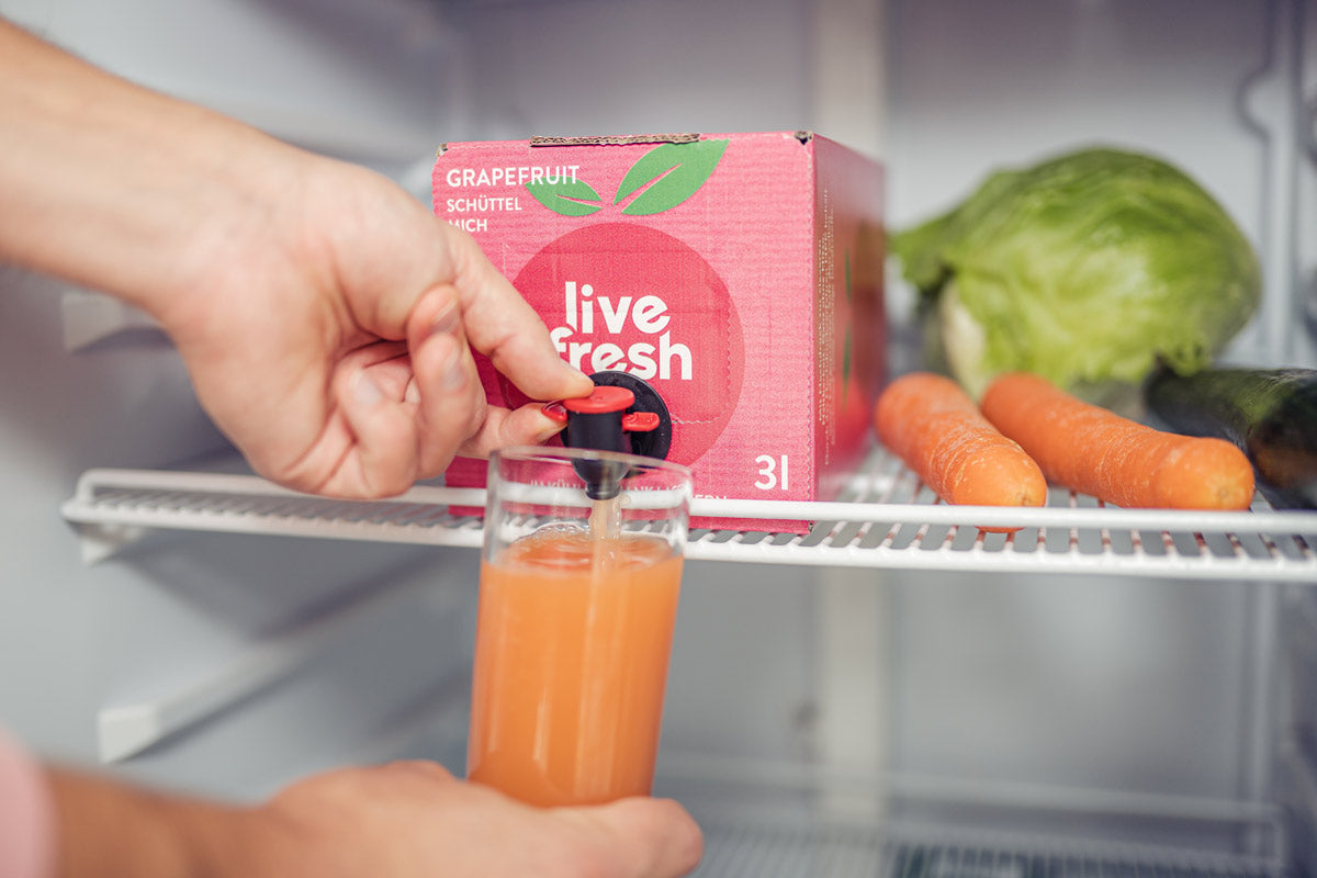 Eine Hand, die in einem Kühlschrank ein Glas mit Grapefruitsaft aus einer rosanen„LiveFresh“ 3-Liter-Box zapft. Auf der Saftbox steht „Grapefruit – Schüttel mich“. Neben der Box liegen frische Karotten, Salat und eine Gurke. Der Zapfhahn der Box wird betätigt, um den frischen Grapefruitsaft ins Glas zu füllen.