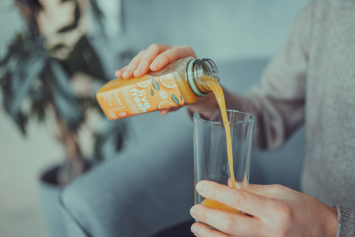 Eine Person gießt Orangensaft aus einer LiveFresh Flasche in ein Glas. Glas und Hand sind scharf, der Oberkörper und der Hintergrund mit Sofa und Pflanze verschwommen.