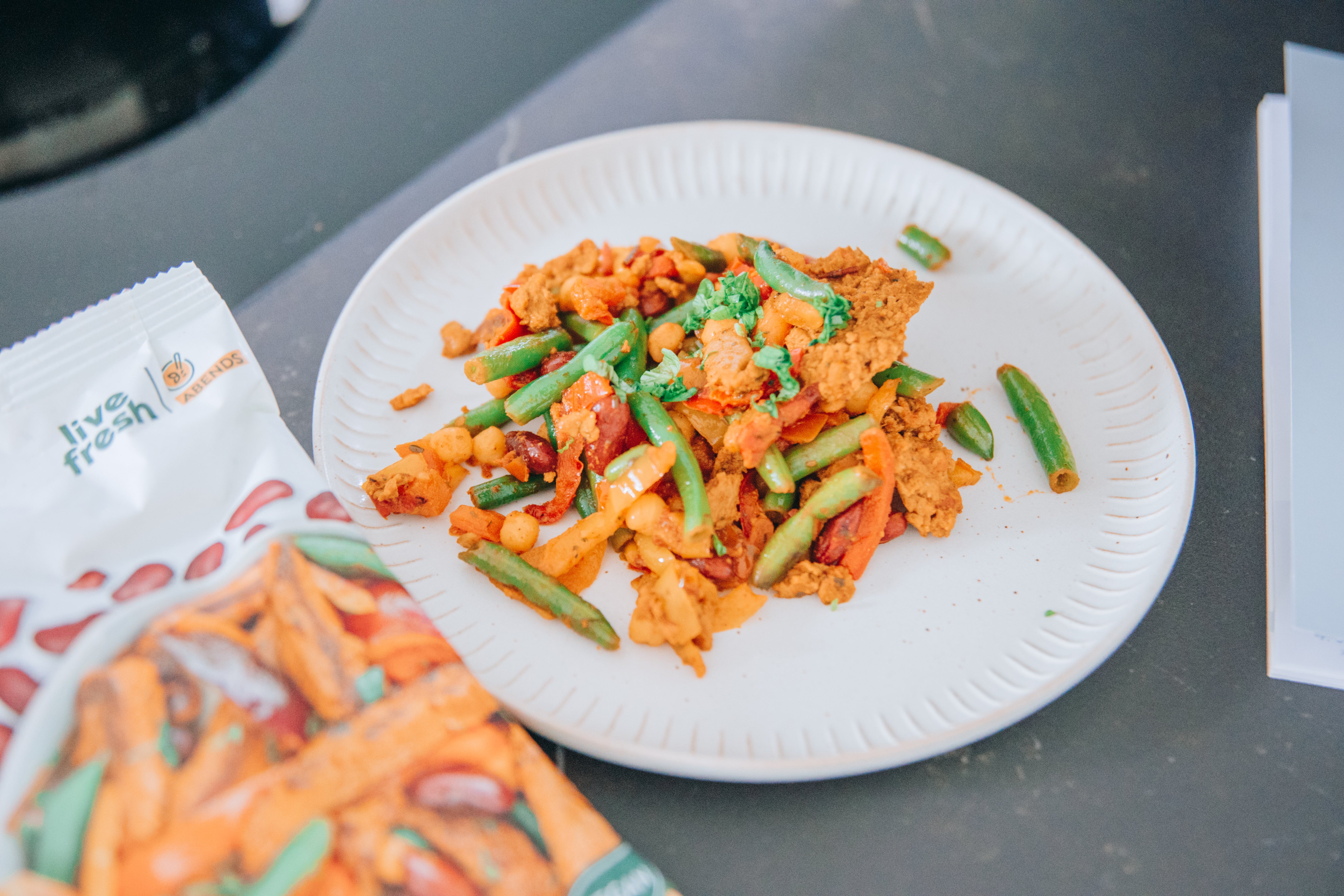 Ein Teller mit Live Fresh Fajita Mexican Style, bestehend aus grünen Bohnen, roten Bohnen, Paprika und Kichererbsen, mit einer Packung der veganen Mahlzeit daneben. Keyword: Wie viel Eiweiß pro Tag.