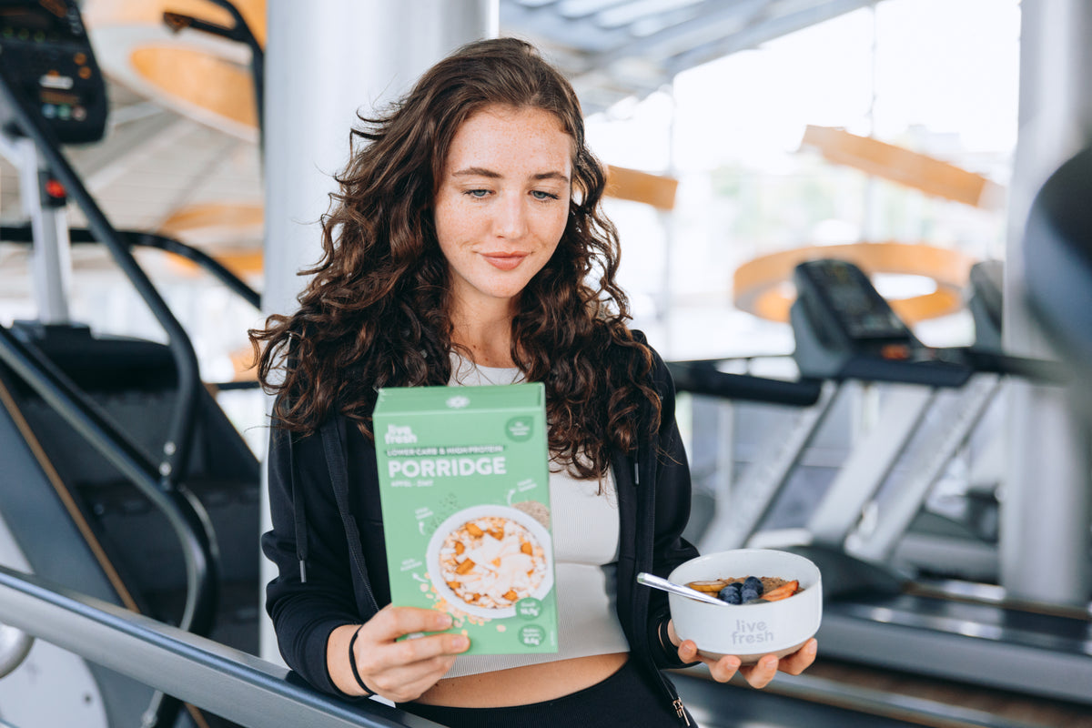 Eine Frau mit lockigem Haar hält eine Packung Live Fresh Porridge und eine Schale mit dem zubereiteten Porridge in der Hand, während sie im Fitnessstudio steht. Keyword: Porridge: Kalorien und Nährwerte im Überblick.