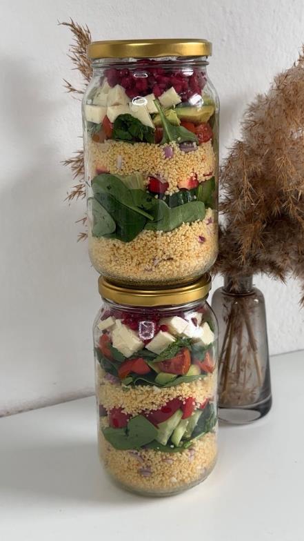 Zwei übereinander gestapelte Einmachgläser mit buntem Couscous-Salat, geschichtet mit Spinat, Tomaten, Gurken, Feta und Granatapfelkernen. Im Hintergrund eine braune Trockenblume in einer Vase auf weißem Untergrund.