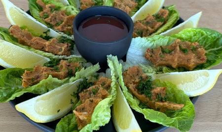 Mercimek Köfte, türkische Linsenbällchen, auf Romanasalatblättern angerichtet, mit Zitronenspalten und einem dunklen Dip in der Mitte auf einem schwarzen Teller serviert.