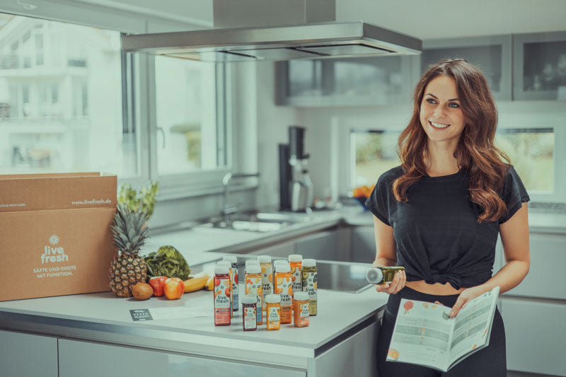 Eine Frau in einer modernen Küche steht neben LiveFresh Säften und einem Informationsblatt. Auf der Arbeitsfläche befinden sich Obst und eine Box mit der Aufschrift 'LiveFresh'.