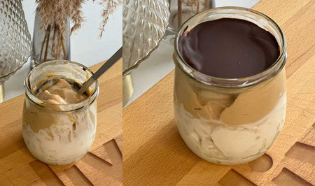 Ein Glas mit Schichten von cremigem Kinder Bueno Dessert und einer Schicht dunkler Schokolade, präsentiert auf einem Holztisch neben dekorativen Glasgefäßen.