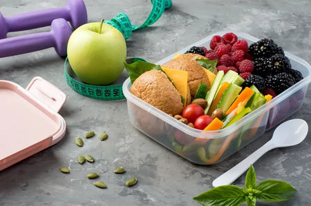 Gesundes Meal-Prep in einer Lunchbox mit Vollkornbrötchen, Käse, Gemüse-Sticks und Beeren, umgeben von Hanteln, Maßband und Apfel – symbolisiert gesunde Ernährung in Verbindung mit Sport und Fasten.