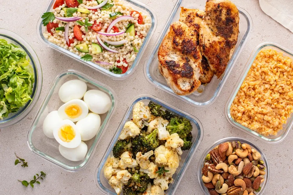 Verschiedene gesunde Mittagsgerichte in Glasbehältern, darunter gegrilltes Hähnchen, Couscous-Salat, gekochte Eier, Brokkoli, Linsen und Nüsse.