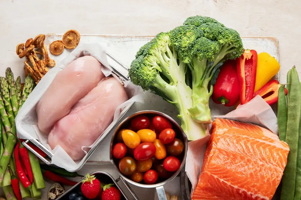 Frische Zutaten für die ketogene Ernährung, darunter Hähnchenbrust, Lachsfilet, Brokkoli, Paprika, Spargel, Pilze und bunte Tomaten, arrangiert auf hellem Untergrund.