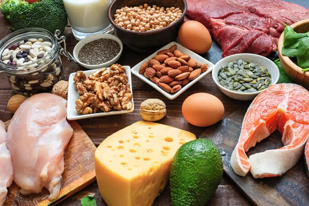 Auswahl natürlicher proteinreicher Lebensmittel wie Lachs, Fleisch, Eier, Käse, Hülsenfrüchte, Nüsse, Samen und Avocado auf einem Holztisch.