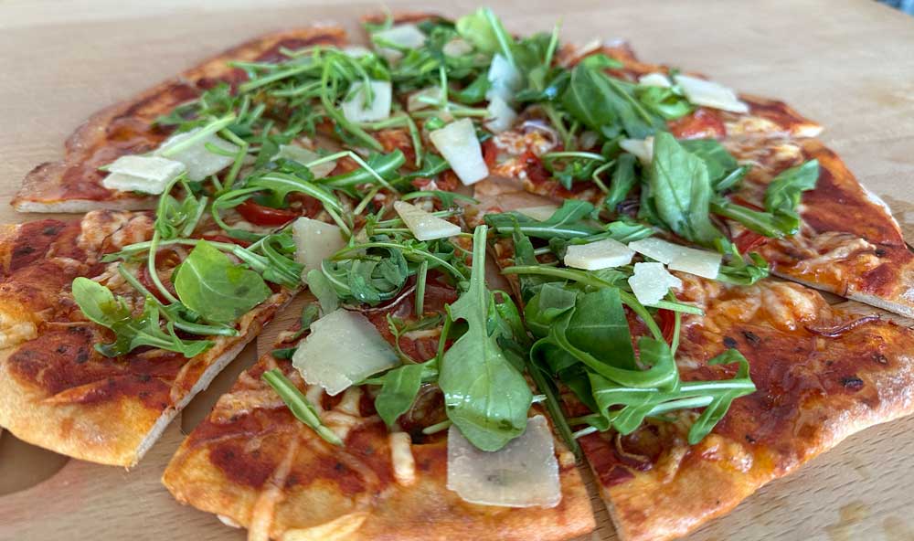 Low Carb Pizza belegt mit Tomatensauce, geschmolzenem Käse, frischem Rucola und Parmesanspänen auf einem hölzernen Untergrund.