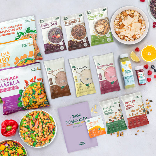 Übersicht der Produkte aus der „7 Tage Food Kur“ von Live Fresh. Zu sehen sind verschiedene Porridge-Sorten, darunter Apfel-Zimt, Schoko und Beere-Vanille, sowie gefrorene vegane Mahlzeiten wie „Kichererbsen Marokkanische Art“, „Kurkuma Curry“ und „Vegan Tikka Masala“. Daneben gibt es vegane Trinkmahlzeiten in verschiedenen Geschmacksrichtungen, Protein-Snacks und Ergänzungsmittel wie die „Tagesheld“-Kapseln. Auch Getränke wie der „Happy Lemon“-Saft und ein Beeren-Shot sind abgebildet.