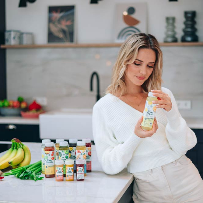 A young woman stands in the kitchen with juices from LiveFresh next to him and a Happy Lemon Juice in your hand. 