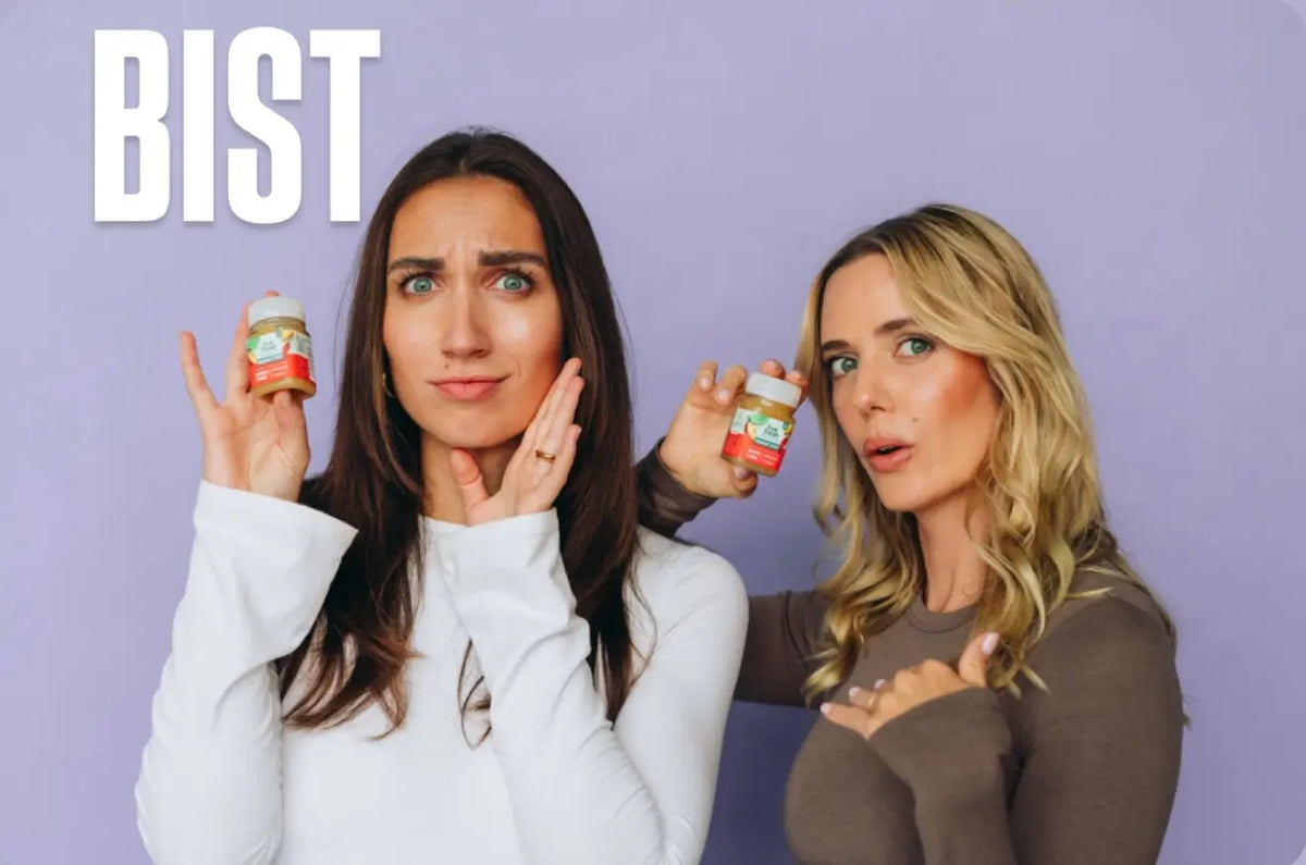 Zwei Frauen zeigen kleine Fläschchen mit Liv Fresh Immunshots vor lilafarbenem Hintergrund. Die brünette Frau blickt skeptisch in die Kamera und hält eine Hand an ihre Wange, während die blonde Frau mit offenem Mund neben ihr steht. Darüber steht "BIST".