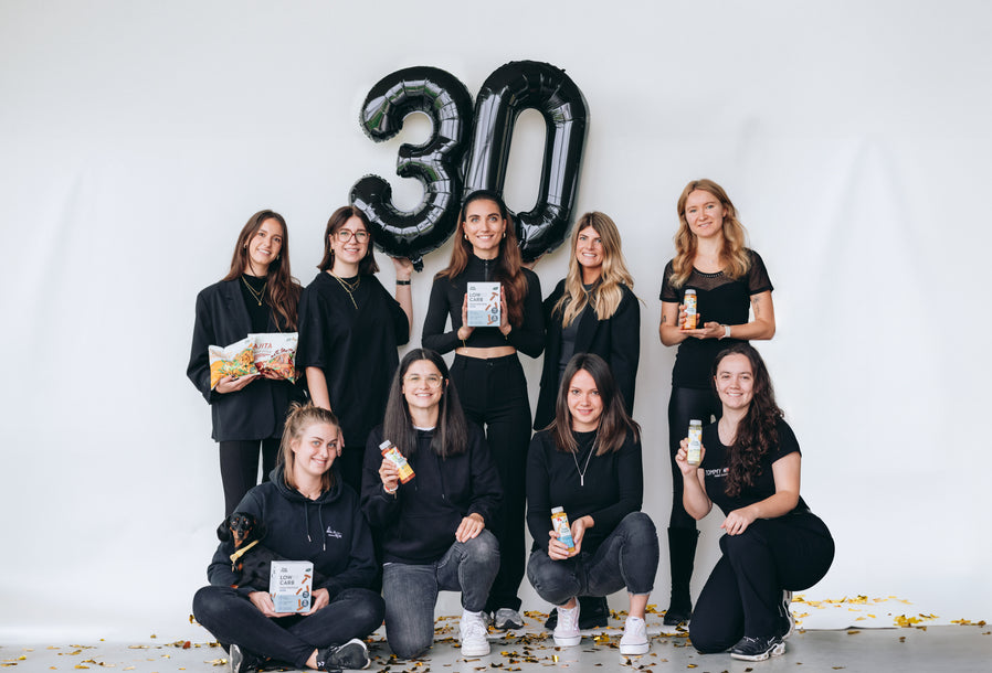 Eine Gruppe von neun Frauen posiert vor einem weißen Hintergrund mit großen, schwarzen Ballons in der Form der Zahl "30". Einige halten Produkte wie Low-Carb-Shakes und Snacks in den Händen, und eine Person hat einen kleinen Hund auf dem Schoß. Am Boden sind goldene Konfetti verstreut, was eine festliche Stimmung vermittelt.