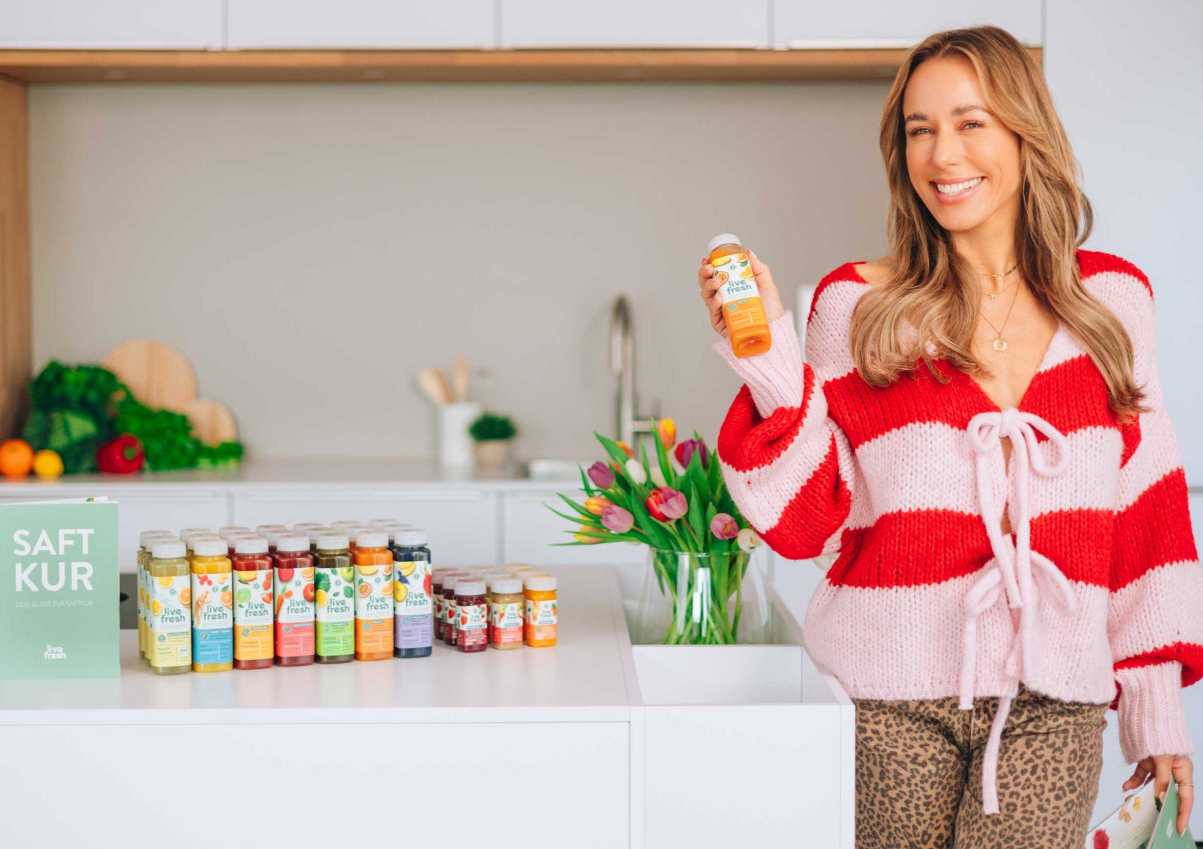 Eine lächelnde Frau mit langen, welligen Haaren trägt einen rot-weiß gestreiften Strickpullover mit Schleifen und hält eine Flasche „Live Fresh“-Saft in der Hand. Sie steht in einer modernen Küche mit einer Reihe bunter Säfte vor sich, neben einem grünen Schild mit der Aufschrift „SAFT KUR“. Auf der Arbeitsplatte steht ein Strauß frischer Tulpen.