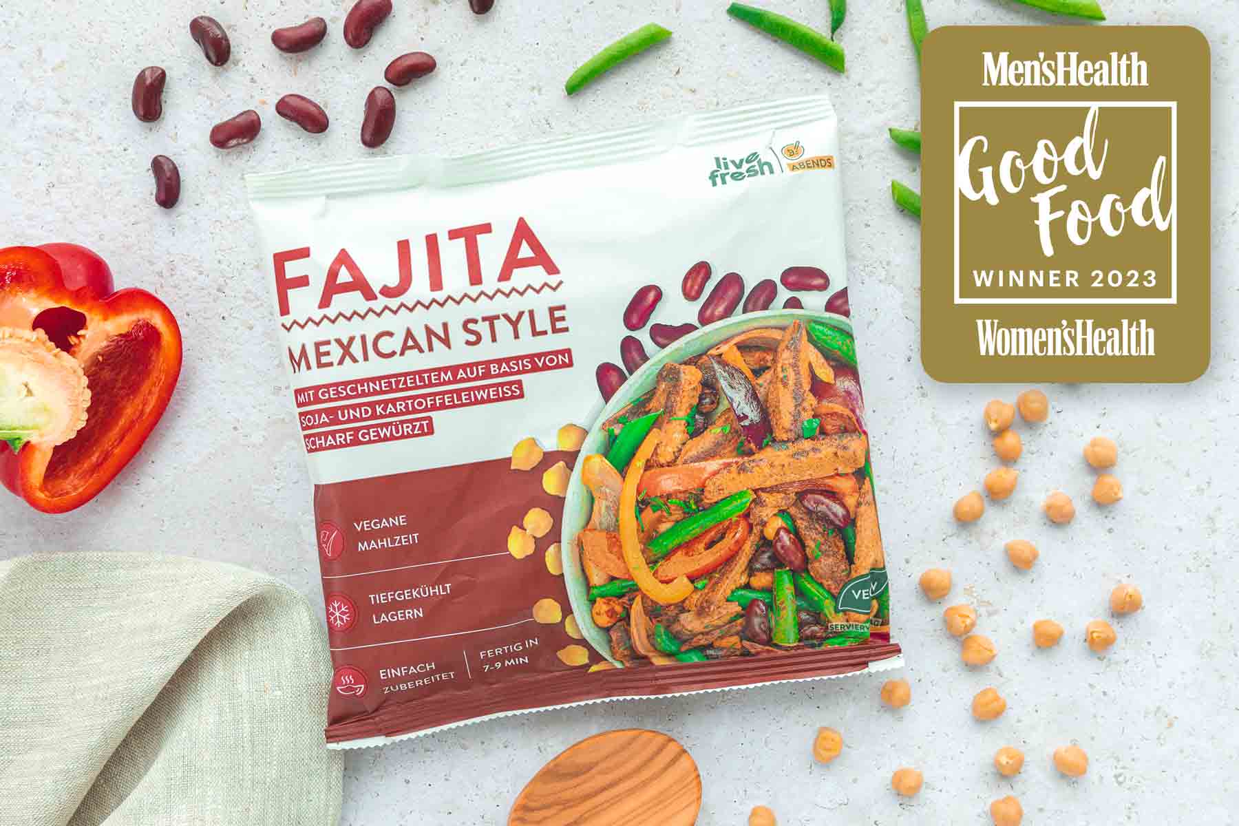 Zeigt eine Packung „Live Fresh Fajita Mexican Style“, die als „Good Food Winner 2023“ von Men's Health und Women's Health ausgezeichnet wurde. Die vegane Mahlzeit enthält geschnetzeltes Soja- und Kartoffeleiweiß und ist scharf gewürzt. Um die Packung herum sind Zutaten wie Paprika, Bohnen und Kichererbsen dekoriert, die auf die Frische und den mexikanischen Stil der Mahlzeit hinweisen.