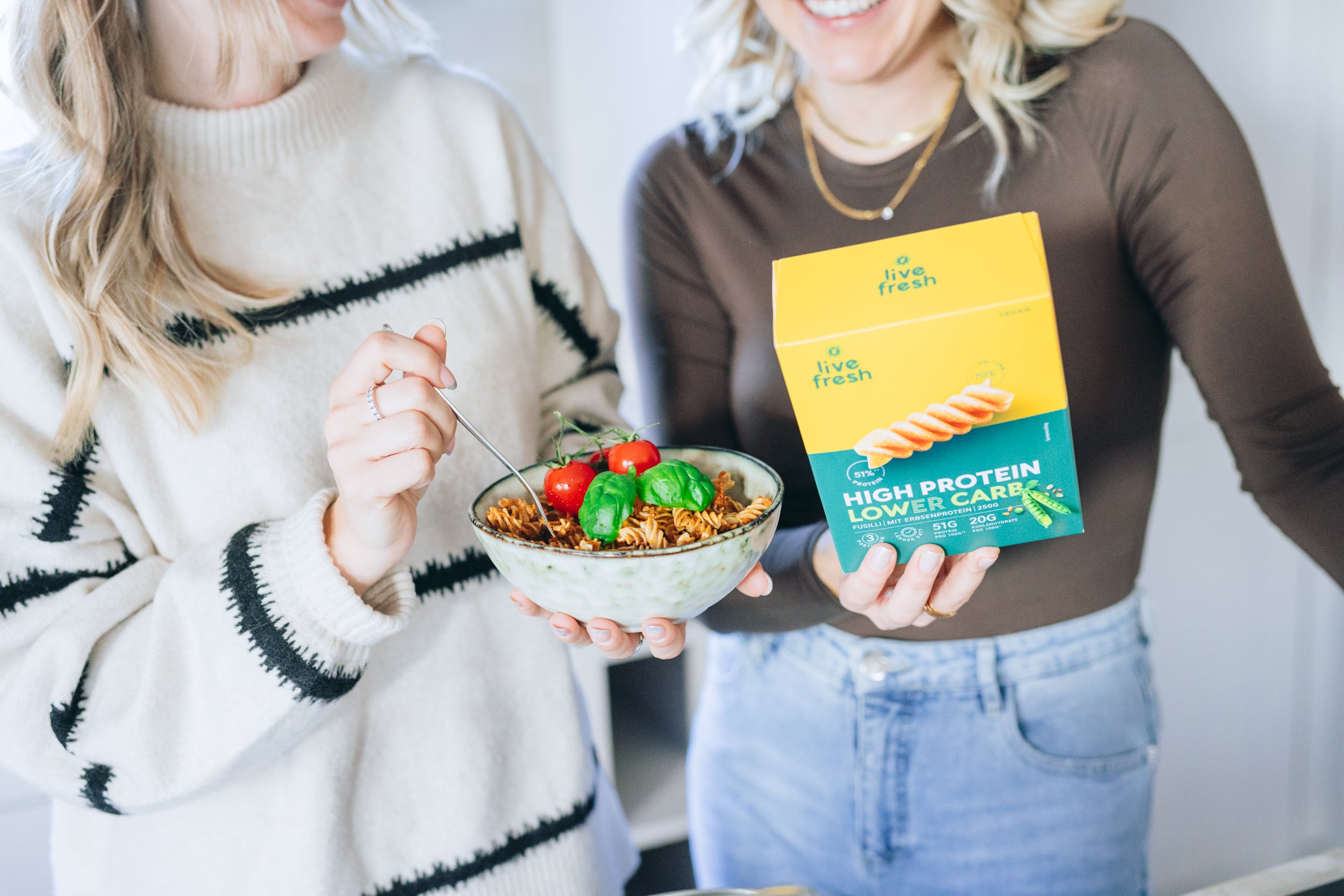 Zwei Frauen, die High Protein und Lower Carb Fusilli von LiveFresh zubereitet haben. Eine Person hält eine Schüssel mit fertigen Nudeln, während die andere die grün-gelbe Verpackung in der Hand hält. 