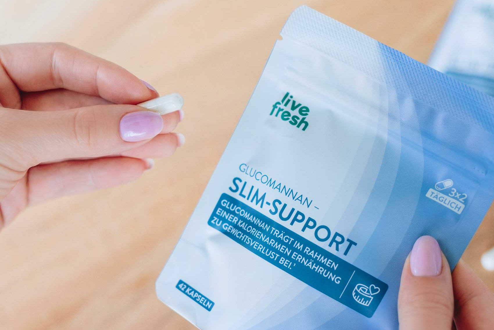 Eine Hand hält eine einzelne Kapsel neben einem Beutel „live fresh Glucomannan Slim-Support“, der teilweise geöffnet ist. Auf der Verpackung steht „GLUCOMANNAN – SLIM-SUPPORT“, „42 KAPSELN“ und „VEGAN“, während die Szene die Einnahme des Produkts im Alltag zeigt.