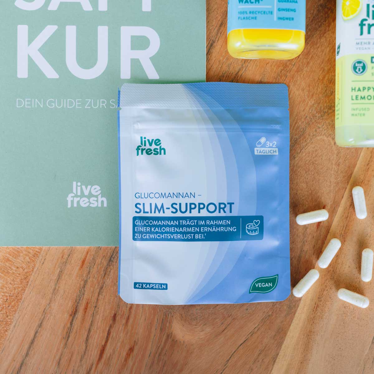Verpackung von „live fresh Glucomannan Slim-Support“ liegt auf einem Holztisch neben verstreuten Kapseln und einem Heft mit der Aufschrift „SAFT KUR“ und „DEIN GUIDE ZUR SAFTKUR“. Auf der Packung steht „GLUCOMANNAN – SLIM-SUPPORT“, „GLUCOMANNAN TRÄGT IM RAHMEN EINER KALORIENARMEN ERNÄHRUNG ZU GEWICHTSVERLUST BEI“, „42 KAPSELN“ und „VEGAN“.