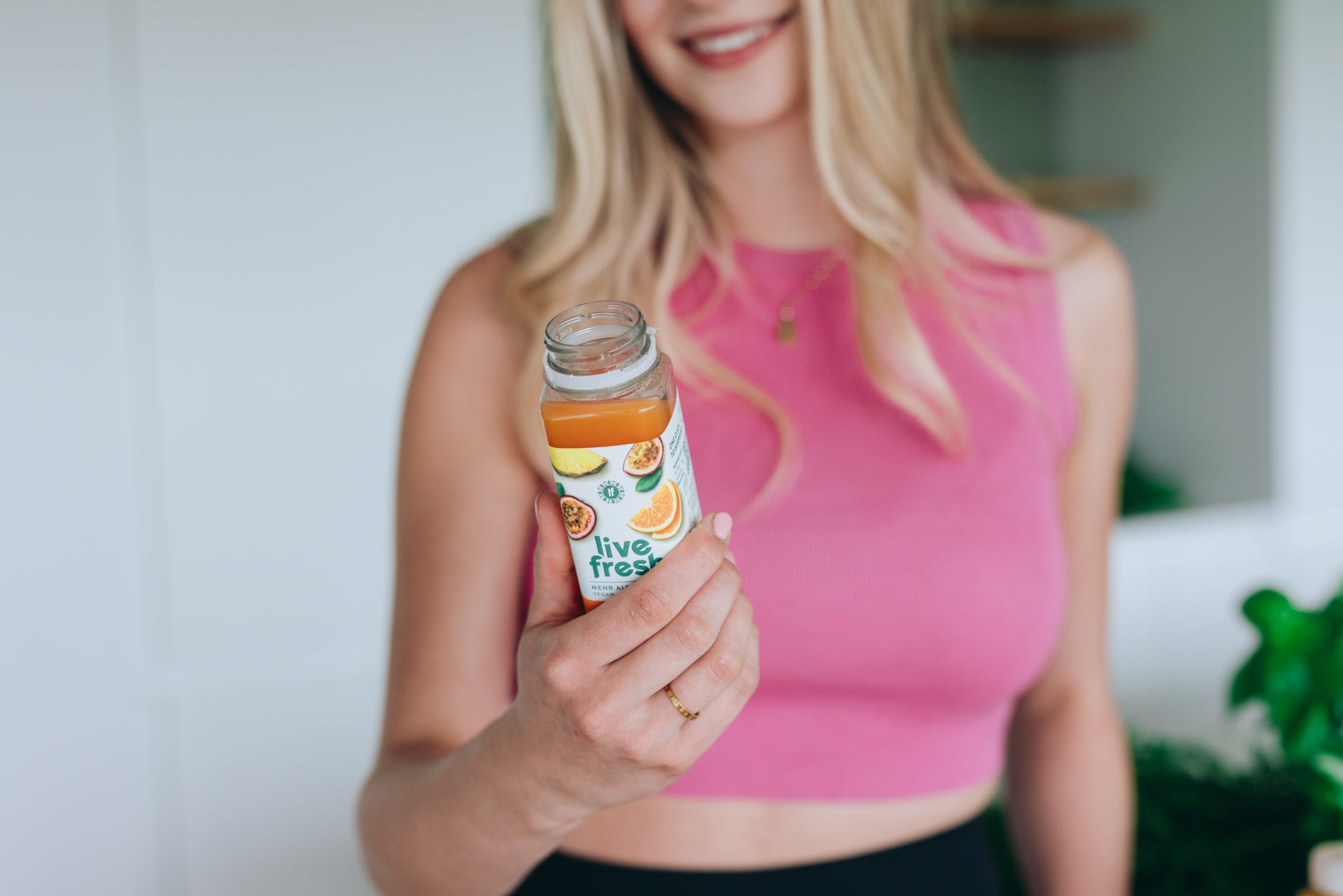 Blonde Frau in pinkem Top hält eine geöffnete Flasche „Live Fresh“ Goldrausch-Saft mit Maracuja, Ananas und Orange auf dem Etikett.