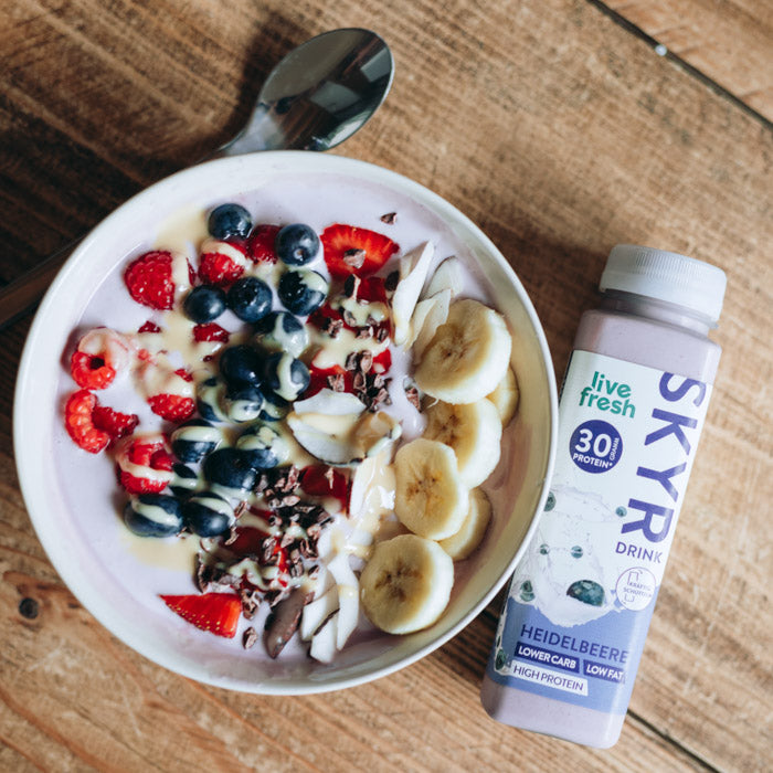 Eine gesunde Bowl, garniert mit Bananenscheiben, Blaubeeren, Erdbeeren, Kokosflocken und Kakaonibs. Daneben steht eine Flasche „Live Fresh Skyr Drink Heidelbeere“ mit 30g Protein. Die Skyr-Flasche wirbt mit einem niedrigen Kohlenhydratgehalt, fettarm und proteinreich, und hebt damit eine ausgewogene, nahrhafte Mahlzeit oder Snack