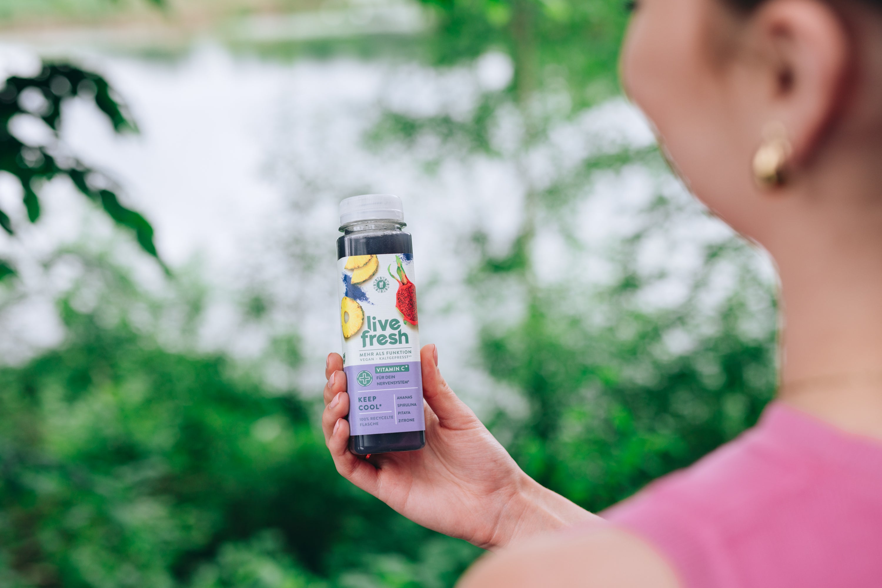 Person im pinken Top hält draußen vor grüner Naturkulisse eine Flasche „Live Fresh Keep Cool“ mit Ananas, Spirulina und Zitrone.